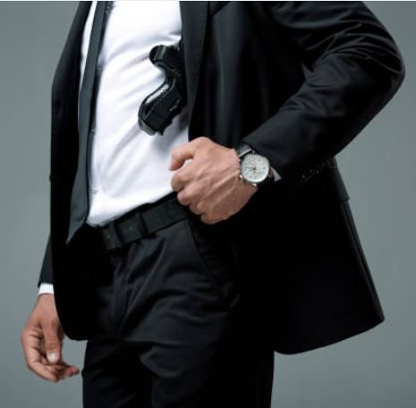 Ein Mann im Anzug mit einer Waffe unter dem Arm und einer Uhr am Handgelenk.