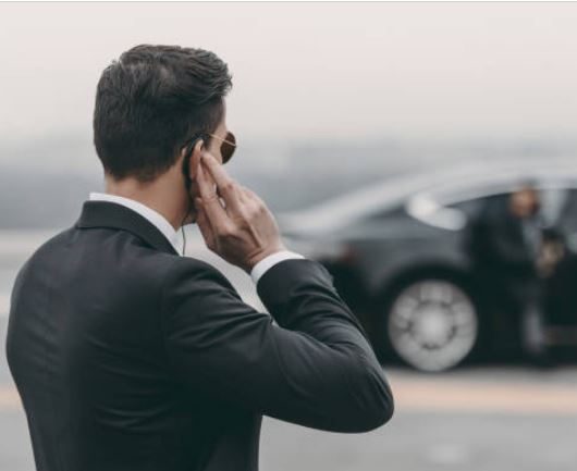 Mann im Anzug mit Sonnenbrille spricht am Telefon neben einem Auto.
